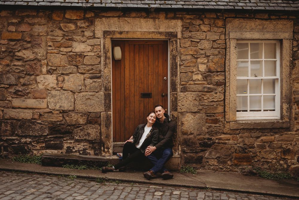 A figure sits alone on stone steps against a rustic brown door and stone wall facade in moody lighting.