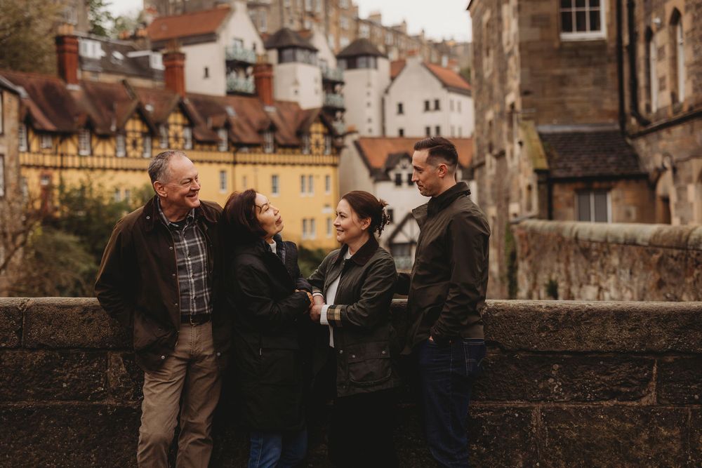 Three people stand together and laugh while chatting outdoors near historic stone buildings on a cloudy day.