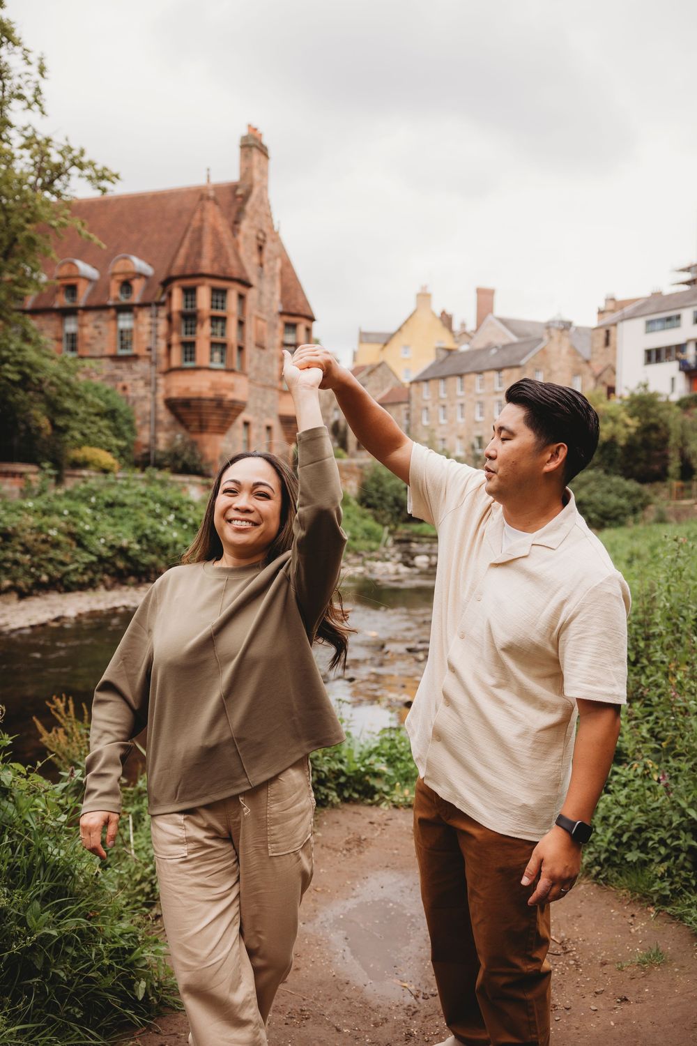 A couple exploring Dean Village in Edinburgh, walking along a path by historic buildings and a stream.