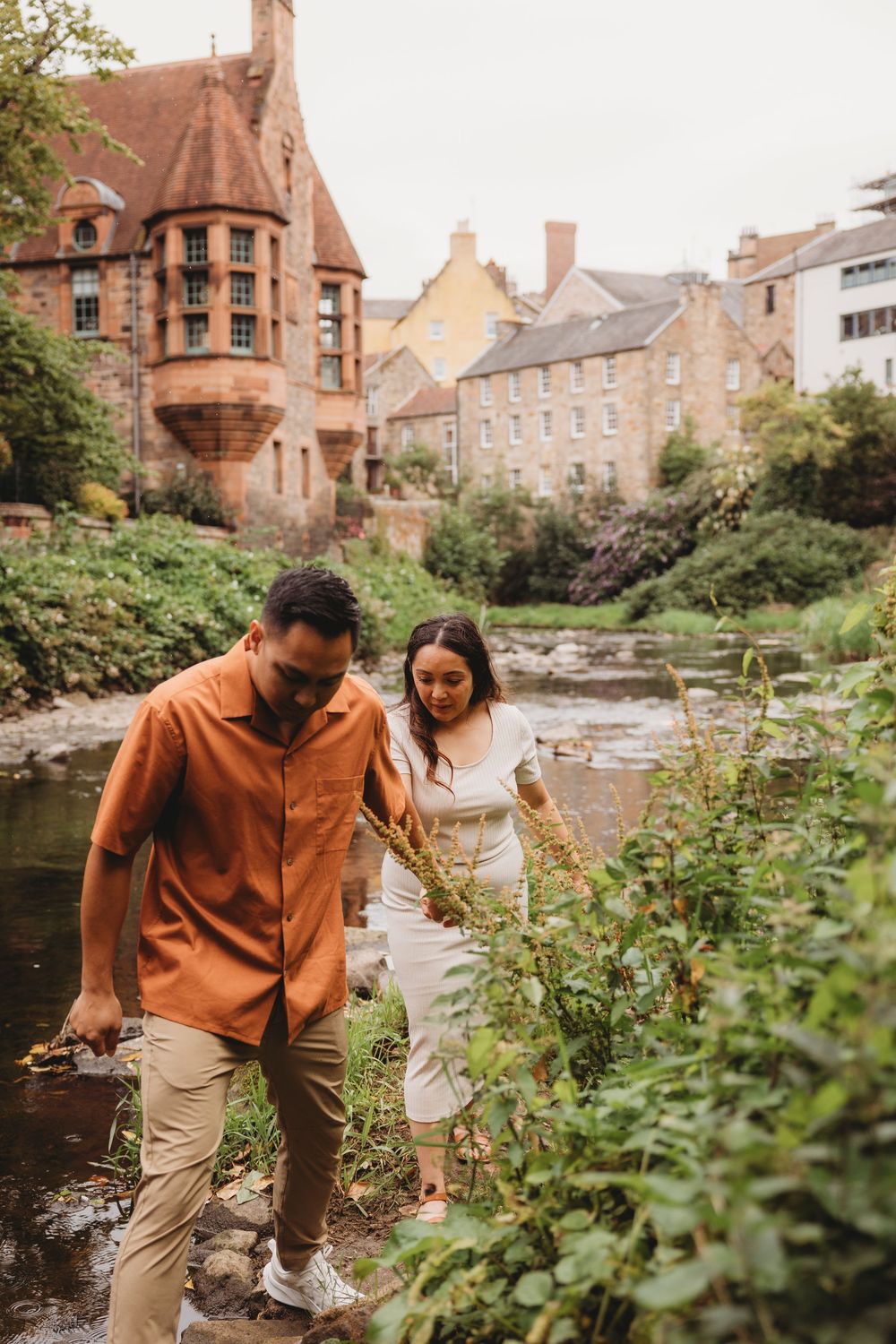 Couple exploring together along a stream beside historic stone buildings and lush gardens in Edinburgh, Scotland.