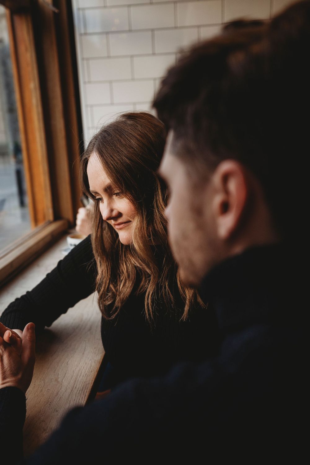 Two people sharing an intimate conversation at a wooden table by a window in warm lighting.