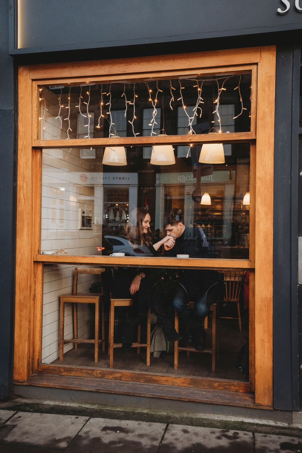View through cafe window showing cozy interior with string lights and pendant lamps above bar seating at night.