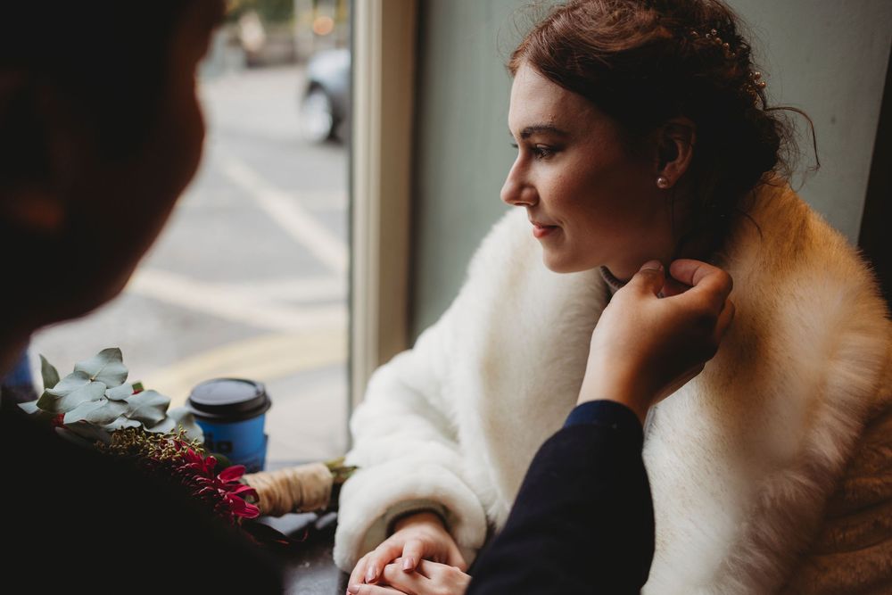 Someone in a white sweater sits thoughtfully by a window in a cafe setting with a blue coffee cup nearby.