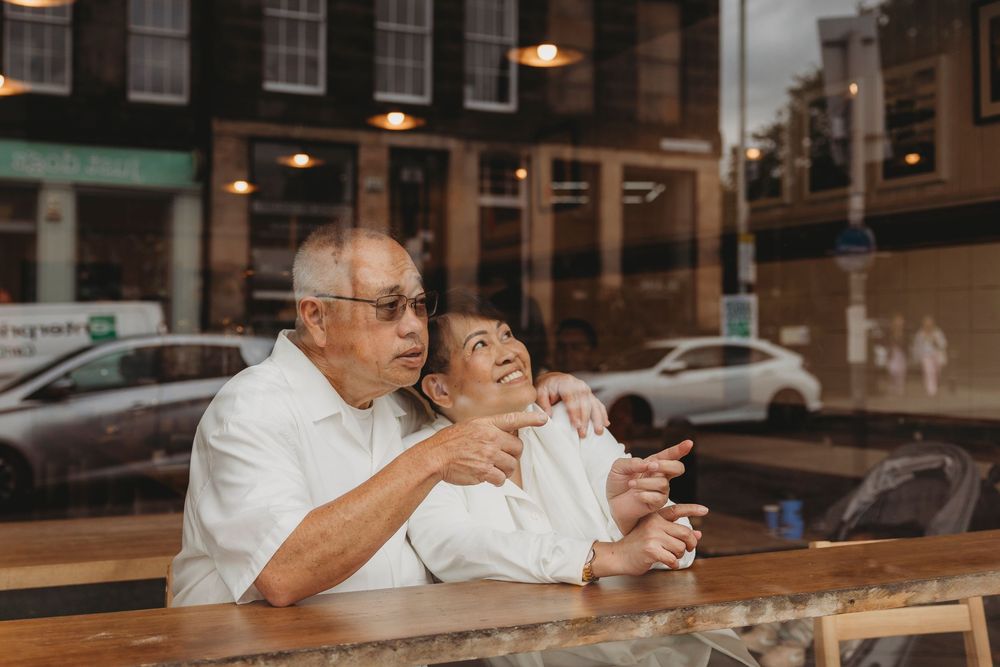 Elderly couple in white shirts laughs together while sitting at wooden counter by window in casual restaurant.