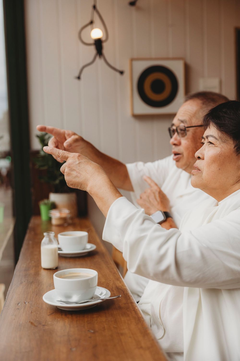 Two seniors in white robes sharing a moment over coffee at a wooden counter with cups and milk on the table.