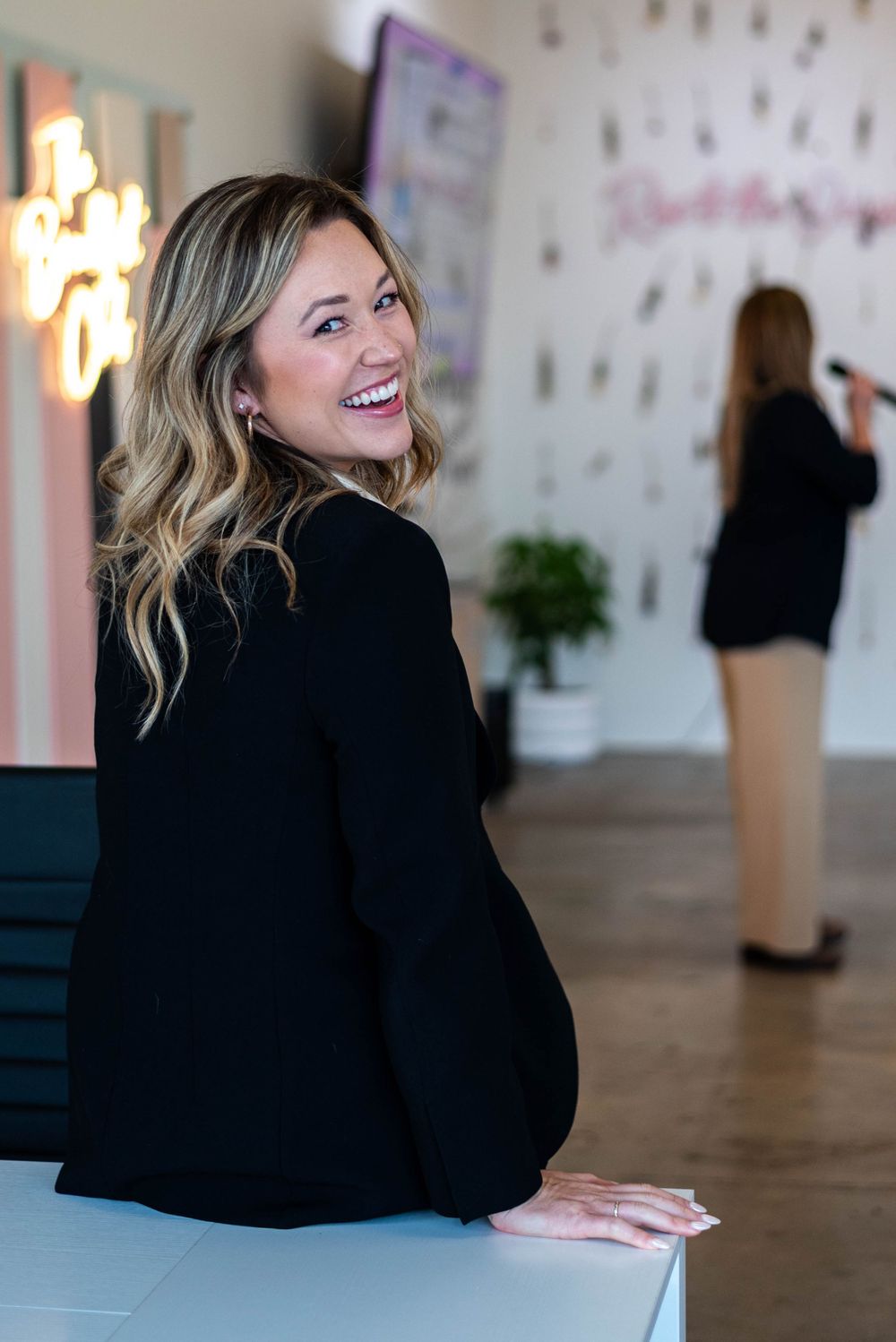 Professional wearing a black blazer smiles while leaning on a counter in a modern office space.