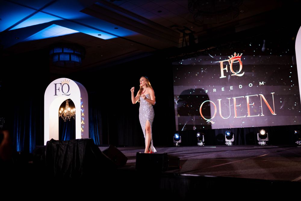 Event presenter on stage with elegant lighting and 'QUEEN' text display behind them.