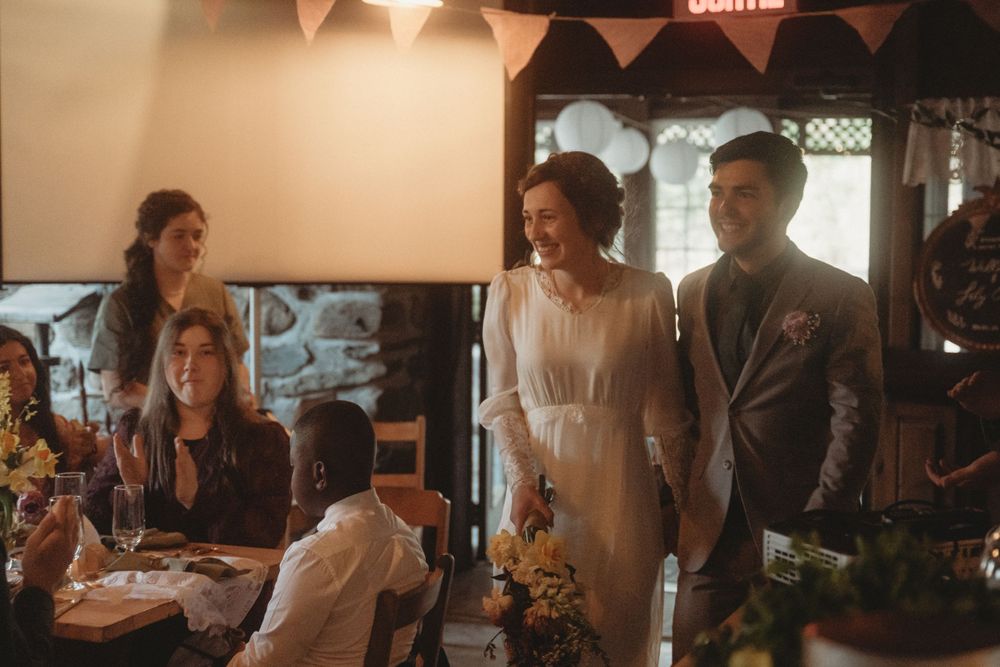 Wedding celebration in a rustic venue with guests gathered around tables under string lights and decorative bunting.