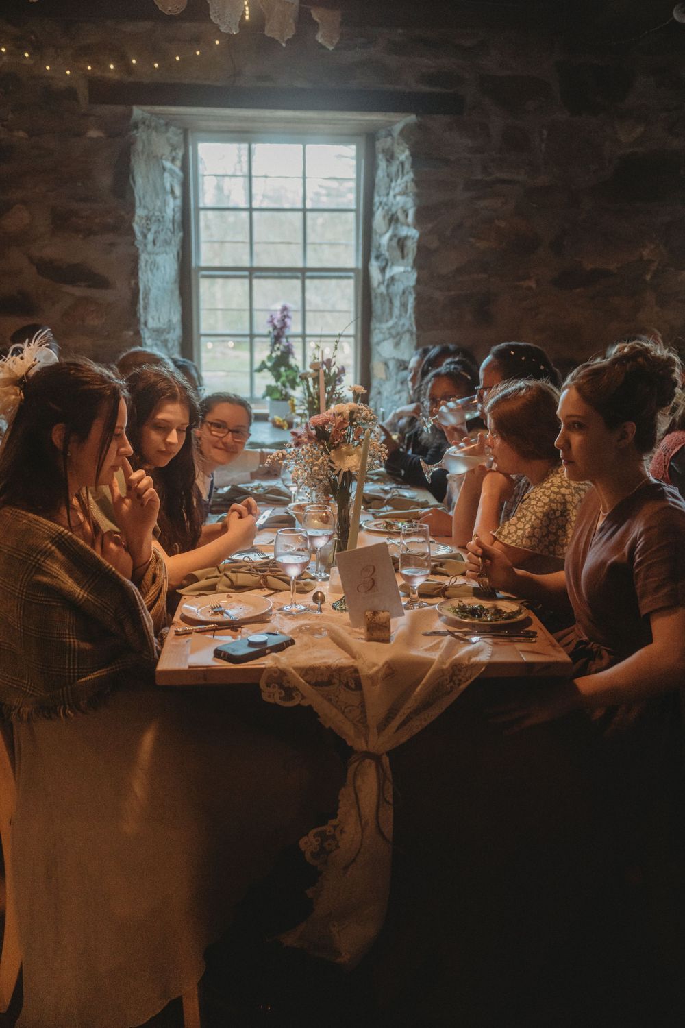 Wedding guests dining at long table near rustic stone window with floral centerpiece and string lights.