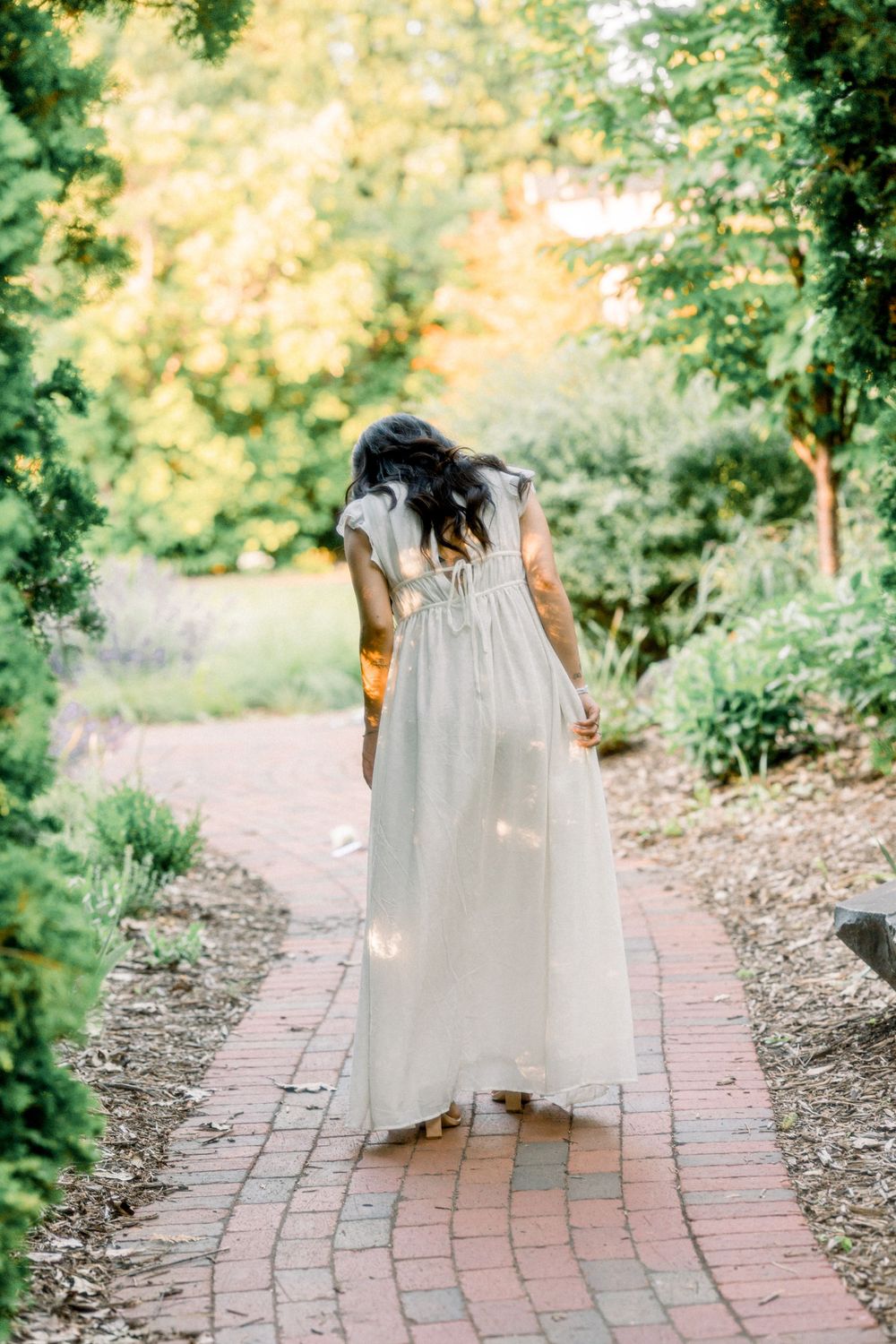 A flowing white dress catches light on a brick path through garden foliage.