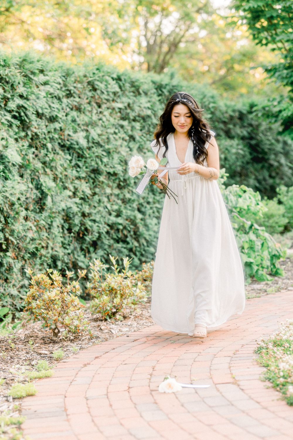 A woman in a flowing white dress walks along a brick garden path holding flowers on a sunny day.