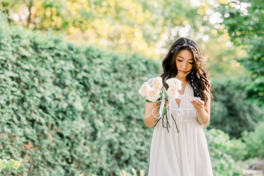 A person in a light colored dress holds flowers while standing in a garden setting.