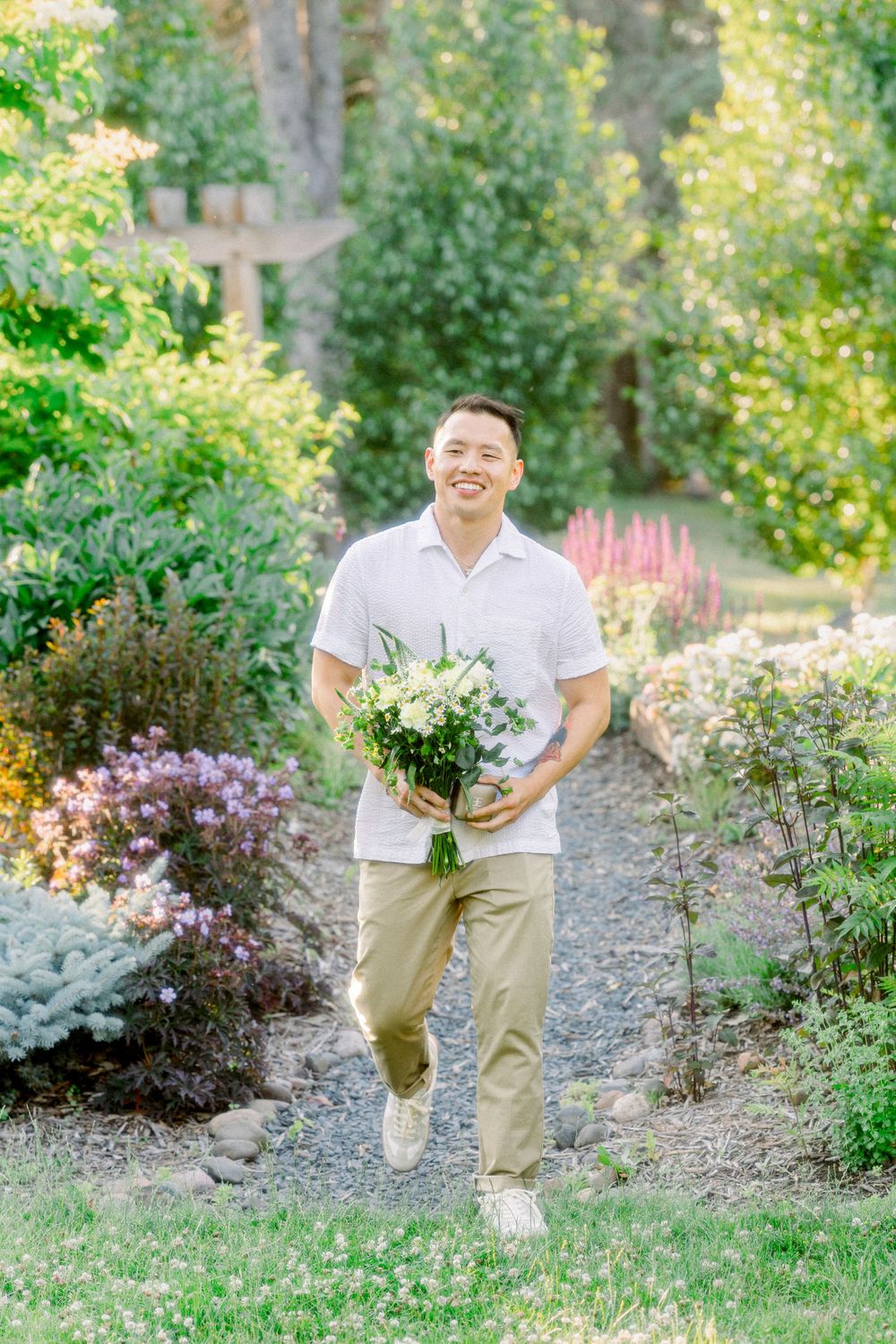 A person carrying a bouquet of white flowers walks along a garden path wearing khaki pants and white shirt.