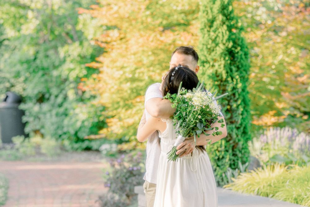 Two people embrace in a garden setting with one holding a bouquet of flowers.