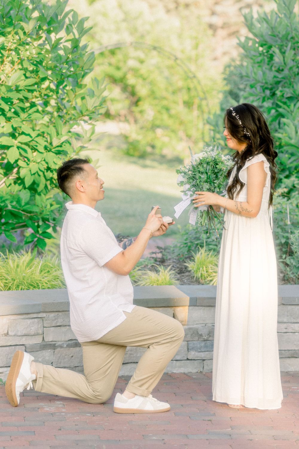 A romantic proposal moment unfolds on a brick pathway in a garden setting.
