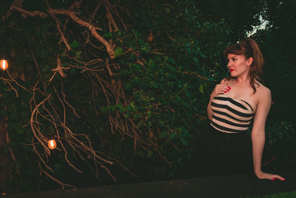 Fashion model in striped swimsuit poses against dark foliage with glowing lights at night.