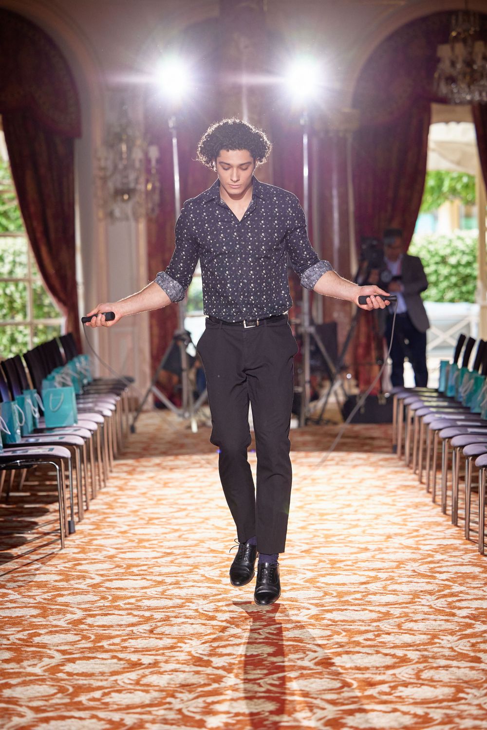 A model walks down an illuminated runway with dramatic lighting and burgundy curtains.