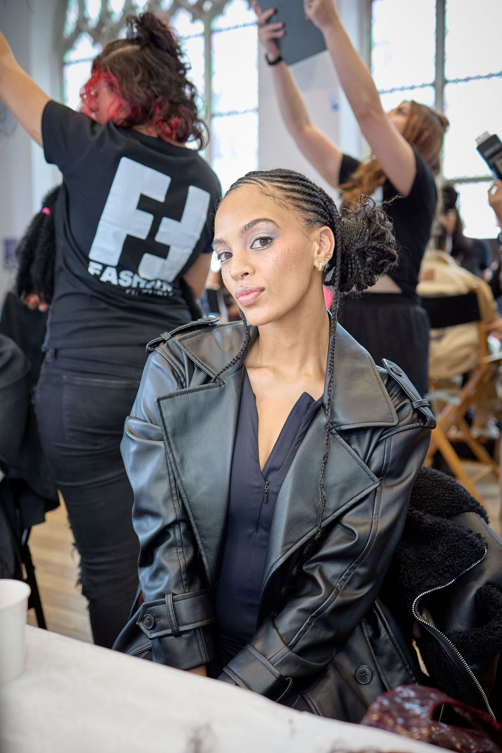Performer wearing a leather jacket poses backstage while others work in the background.