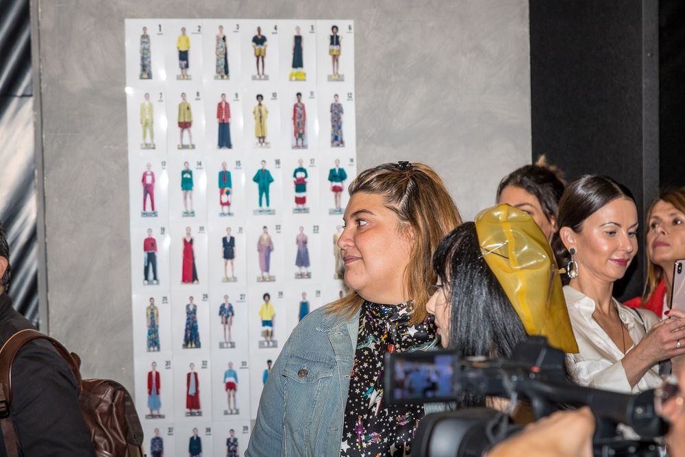 A fashion show preparation area showing a display board with multiple outfit sketches on the wall.