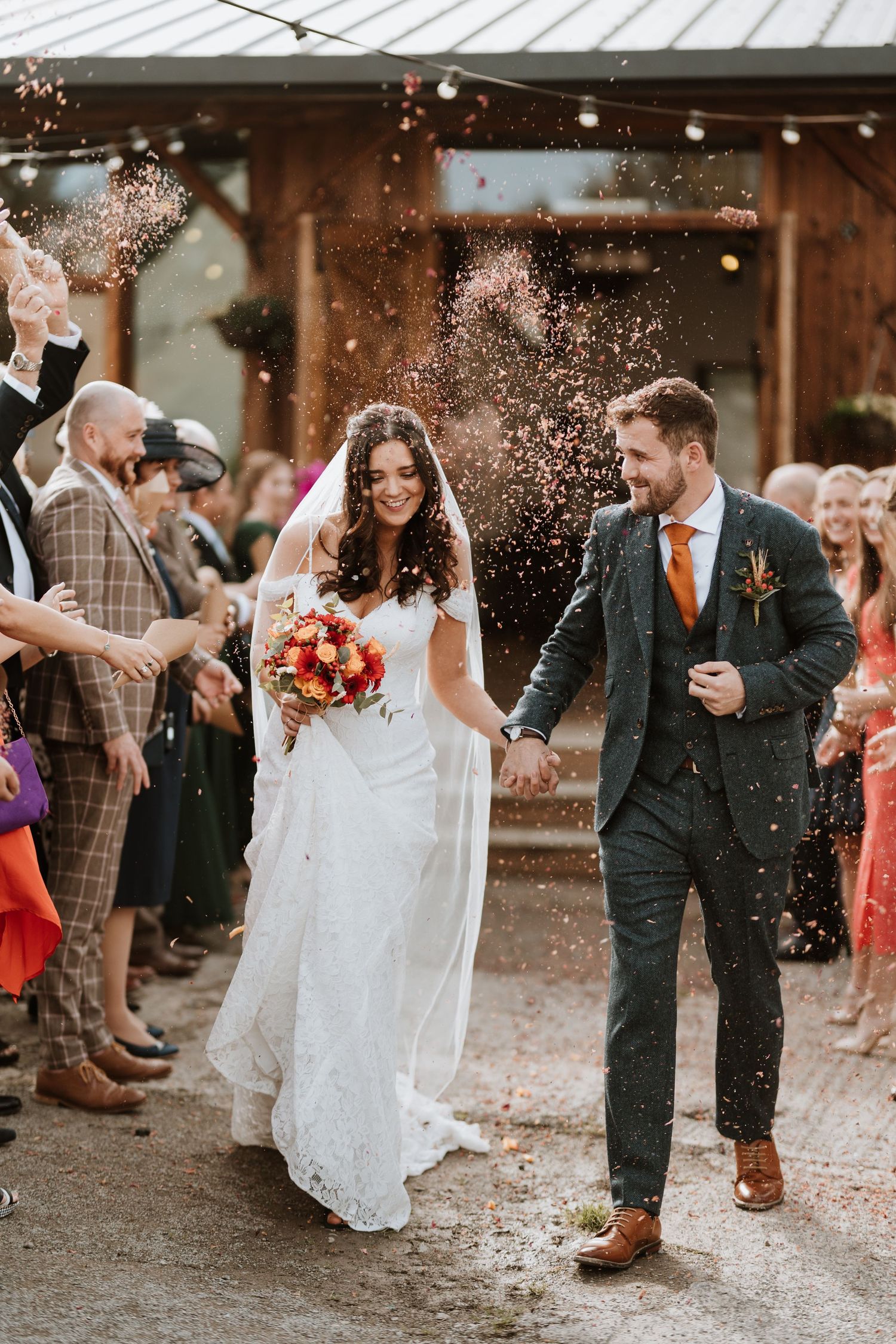 Happy newlyweds walking down aisle while guests throw confetti at rustic barn wedding venue.
