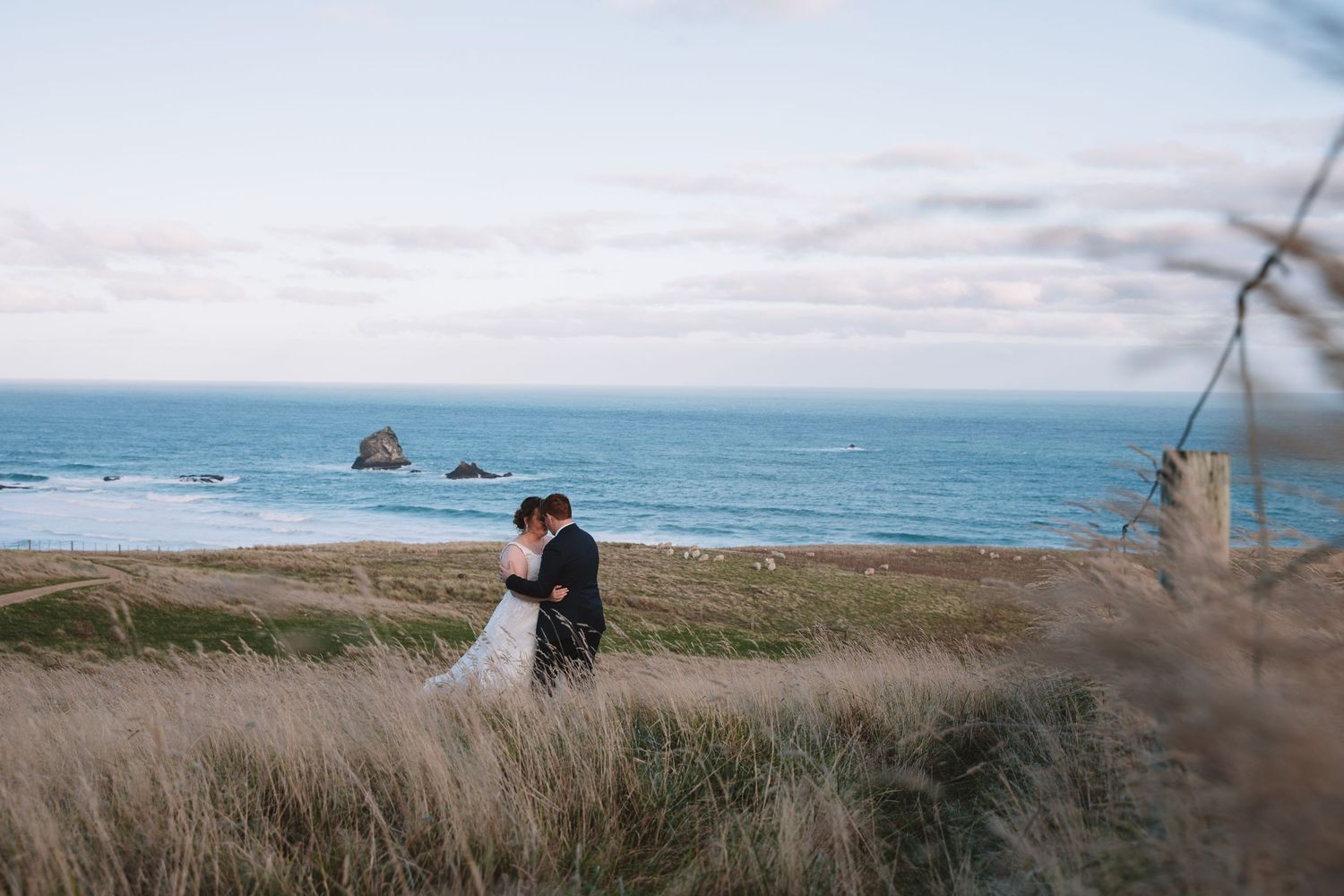 Couple in wedding attire embraces on cliff overlooking rocky ocean coastline.