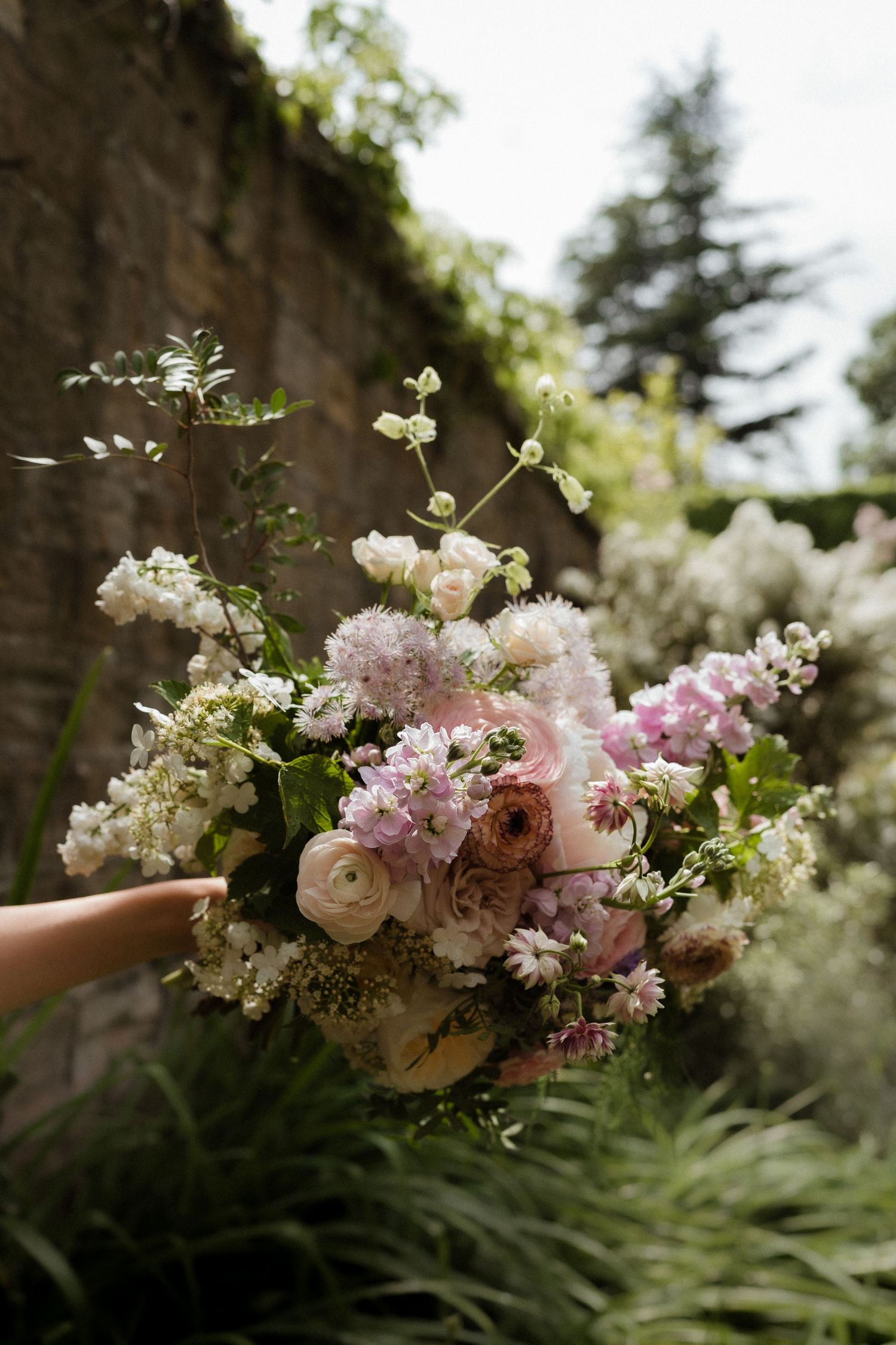 A delicate bouquet of pink and white flowers including ranunculus and cherry blossoms being held up against a garden wall.