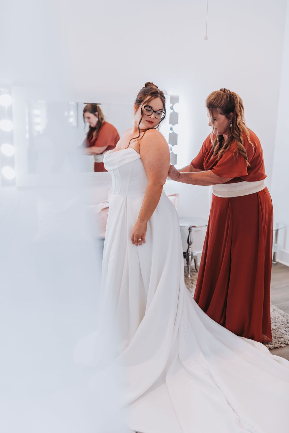 A bride in a white wedding dress and glasses gets fitted by an attendant in a red gown in a bridal suite.
