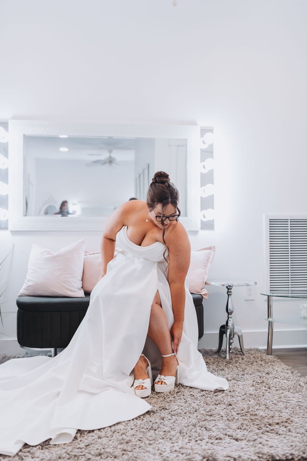 Bride in white wedding gown adjusts her shoe while getting ready in a bedroom with vanity mirror and carpet.