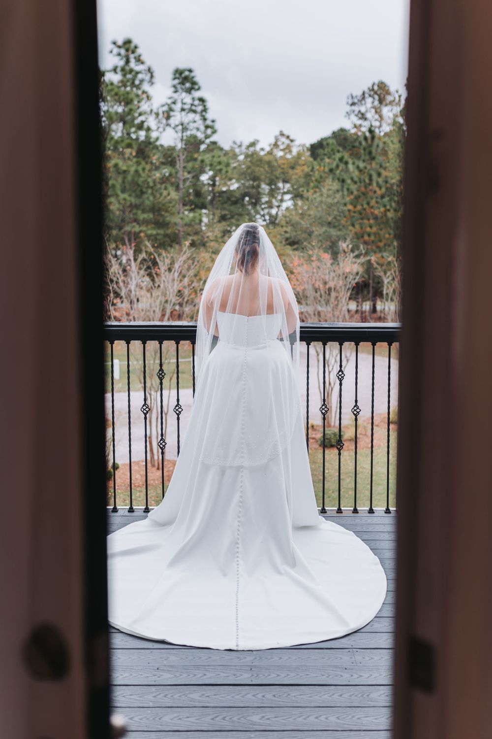 A bride in a white wedding gown with veil stands on a deck overlooking trees and nature in a romantic back-facing pose.