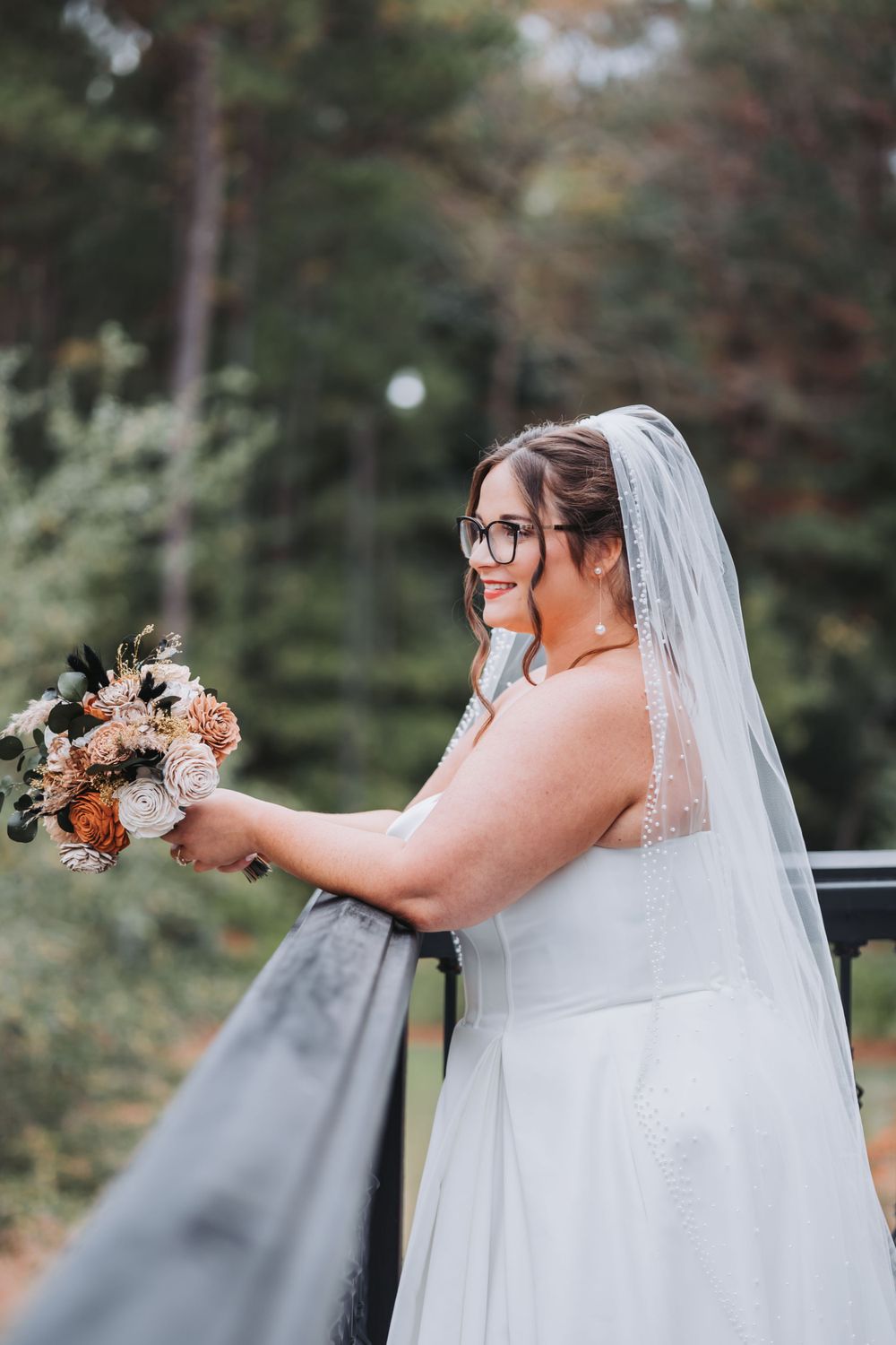A bride in glasses holds an autumn-colored bouquet while standing on a balcony in her white wedding gown.