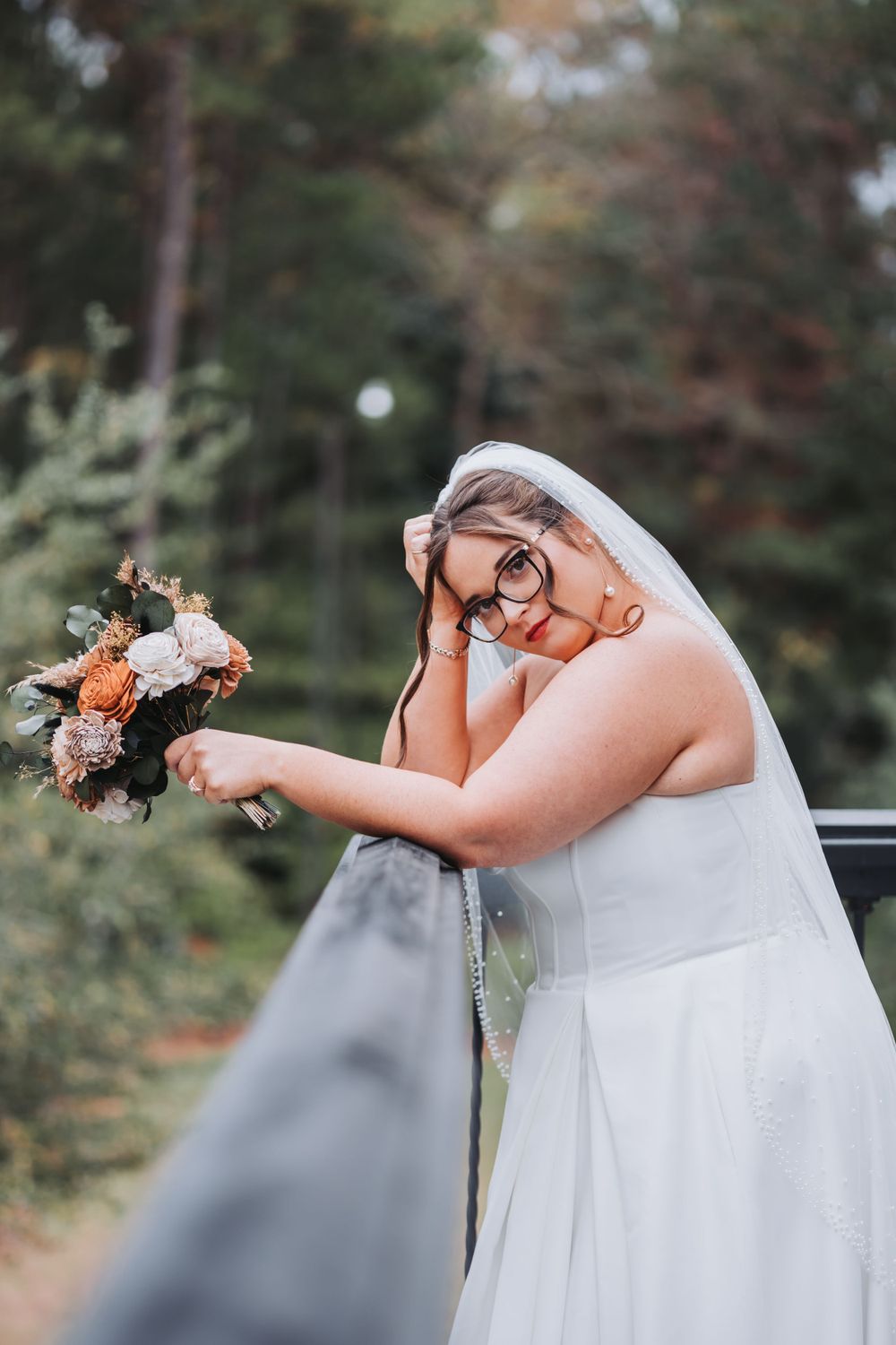 Bride in a white wedding dress and glasses joyfully holds a peach and white floral bouquet outdoors.