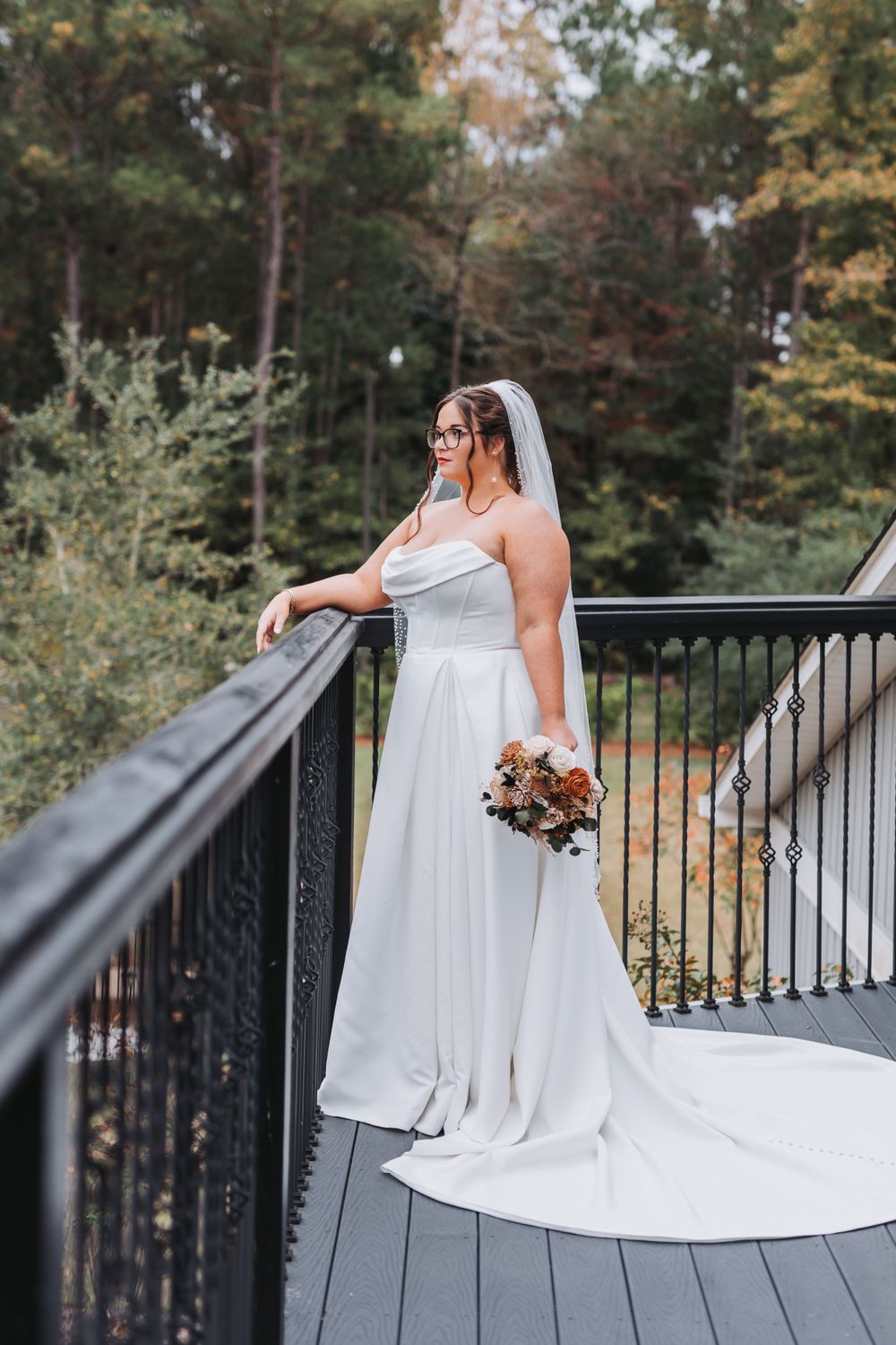 A person in a flowing white wedding dress with train stands elegantly on a wooden deck surrounded by autumn trees.