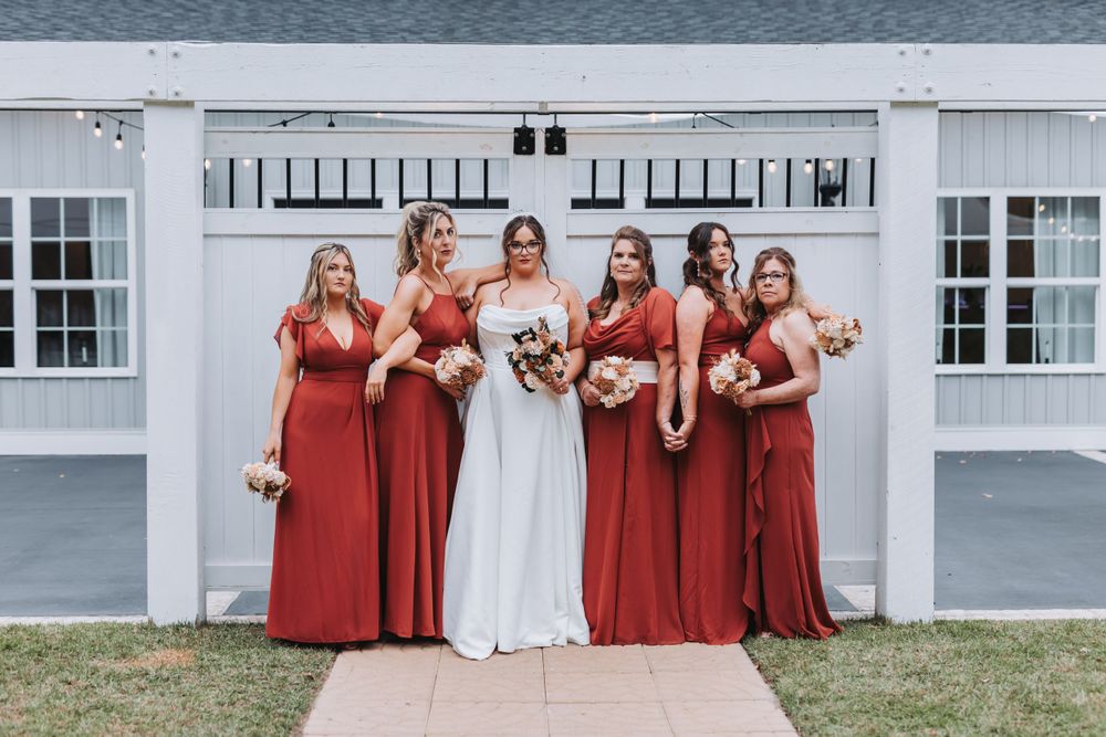 Wedding party with bridesmaids in matching long red dresses posing with bride in white gown against white barn backdrop.