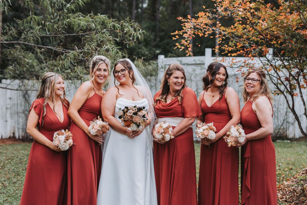 A wedding party poses outdoors in matching burgundy red dresses with autumn foliage in the background.