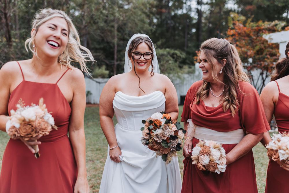 Wedding party in rust-colored bridesmaid dresses pose together outdoors with flowers and laughter.