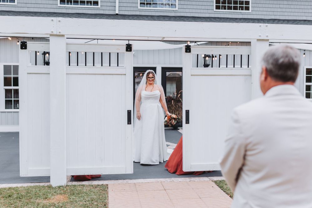 Bride stands in doorway of white barn-style building with rustic door detail.