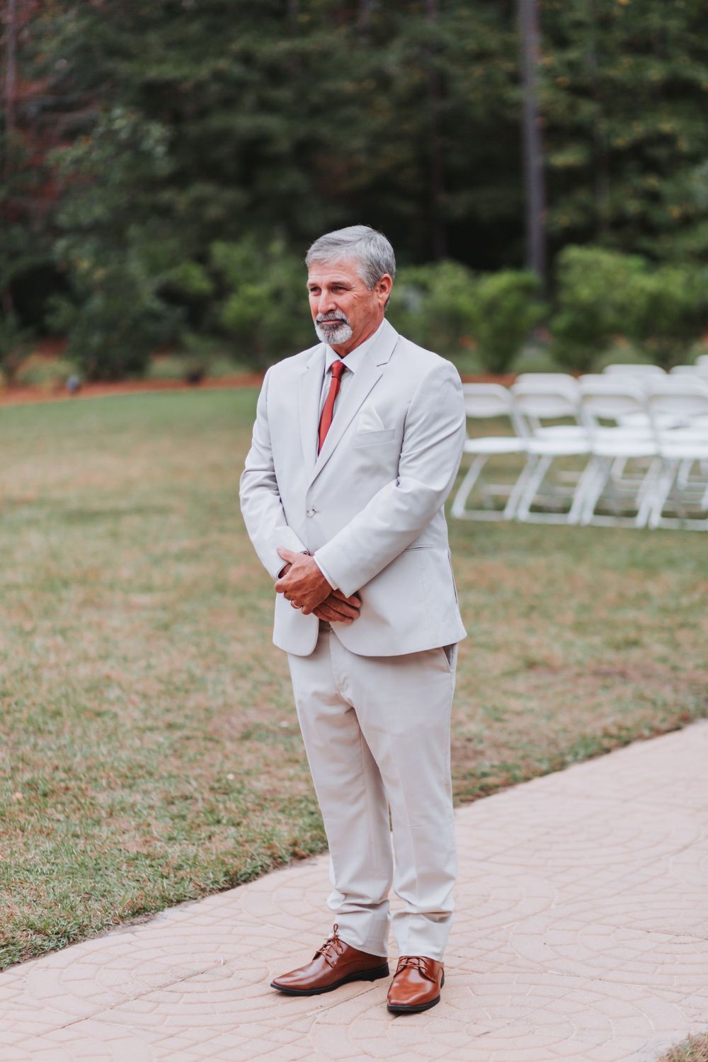 A well-dressed person in a light gray suit and red tie stands on a pathway near rows of white chairs outdoors.