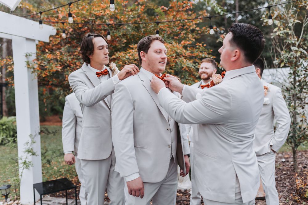 Wedding party members in light gray suits share a lighthearted moment under an outdoor arbor during autumn.