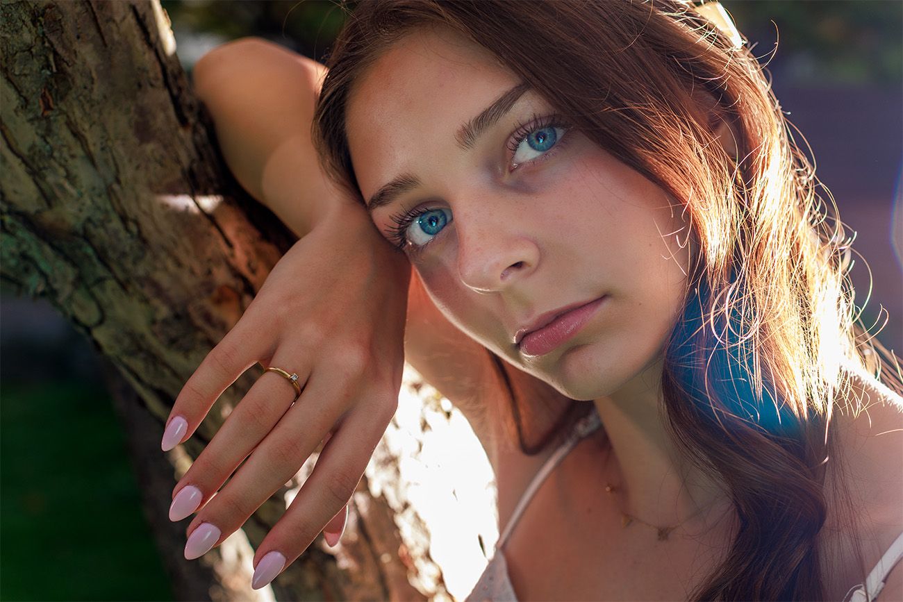 Portrait photo taken outdoors next to a tree trunk with natural sunlight creating a warm glow and artistic lighting effect.