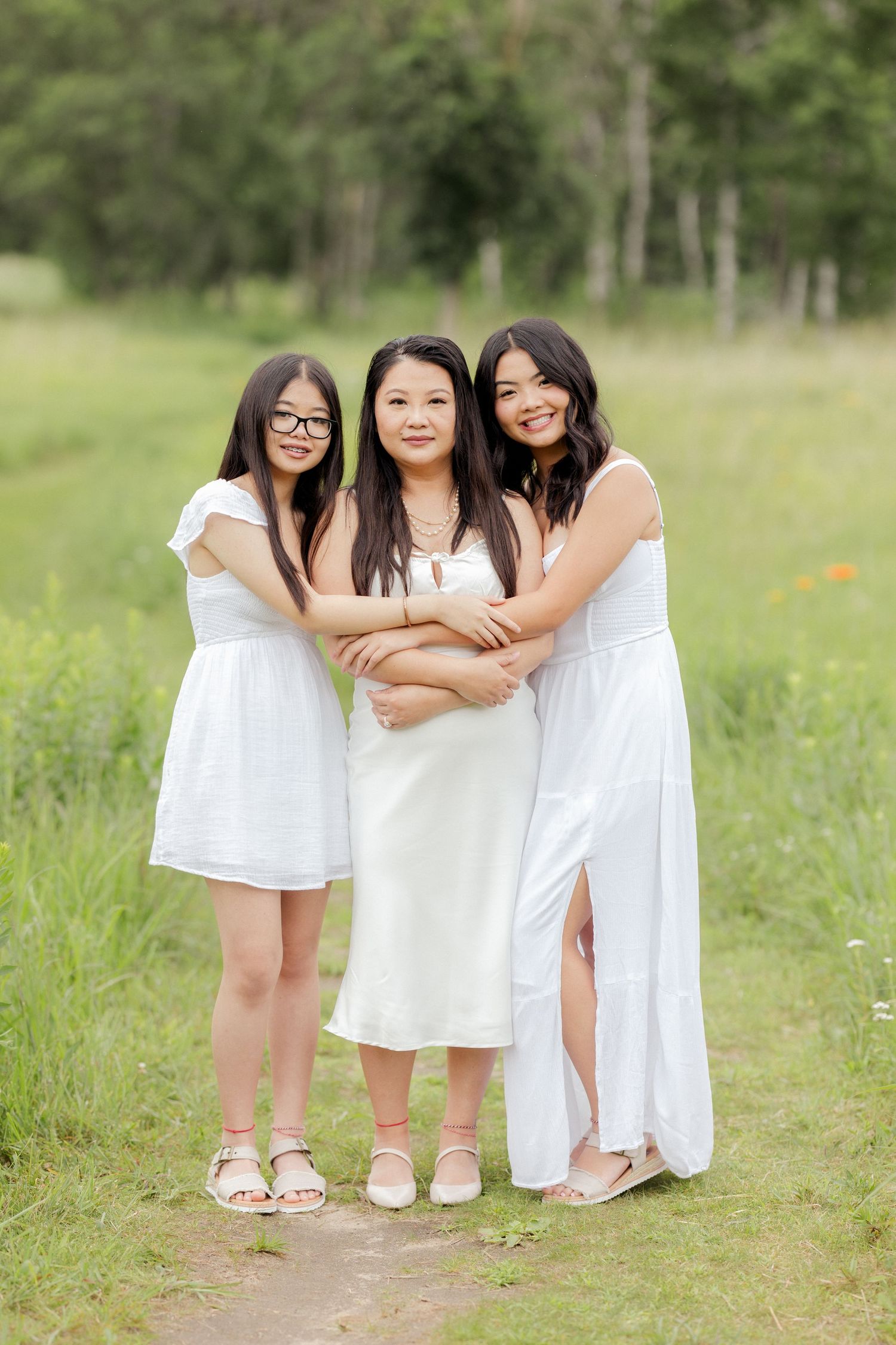 Three women in white dresses embrace while standing on a path through a meadow.