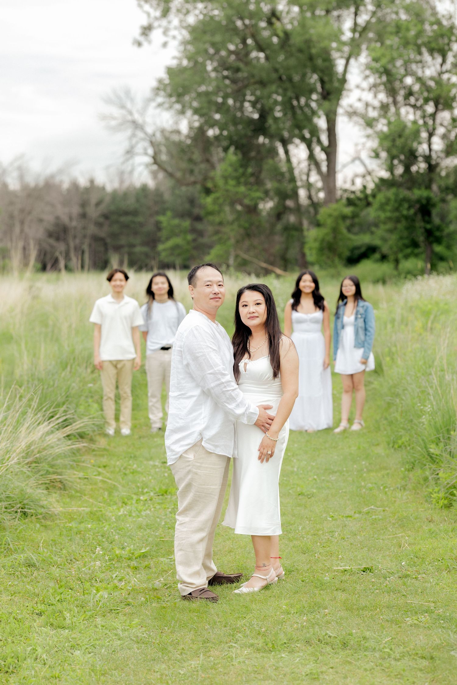 A couple stands in a grassy meadow with friends in light summer outfits gathered behind them.