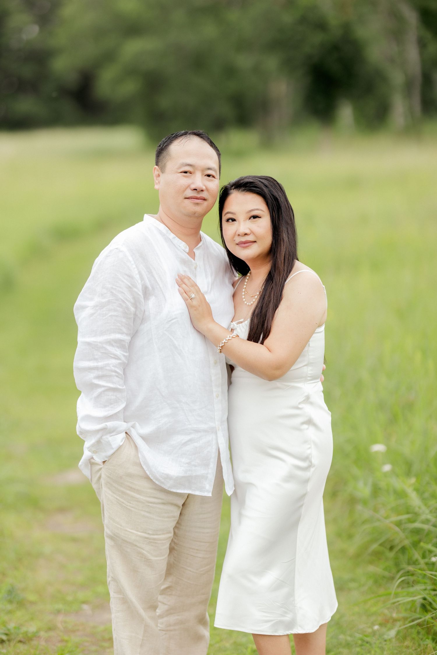 A couple in white and beige clothing pose together in a sunny meadow.