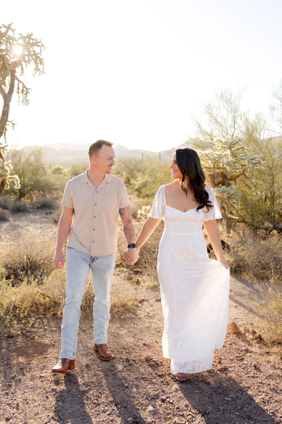 A couple holds hands while walking together in a desert landscape at sunset with desert flora in the background.