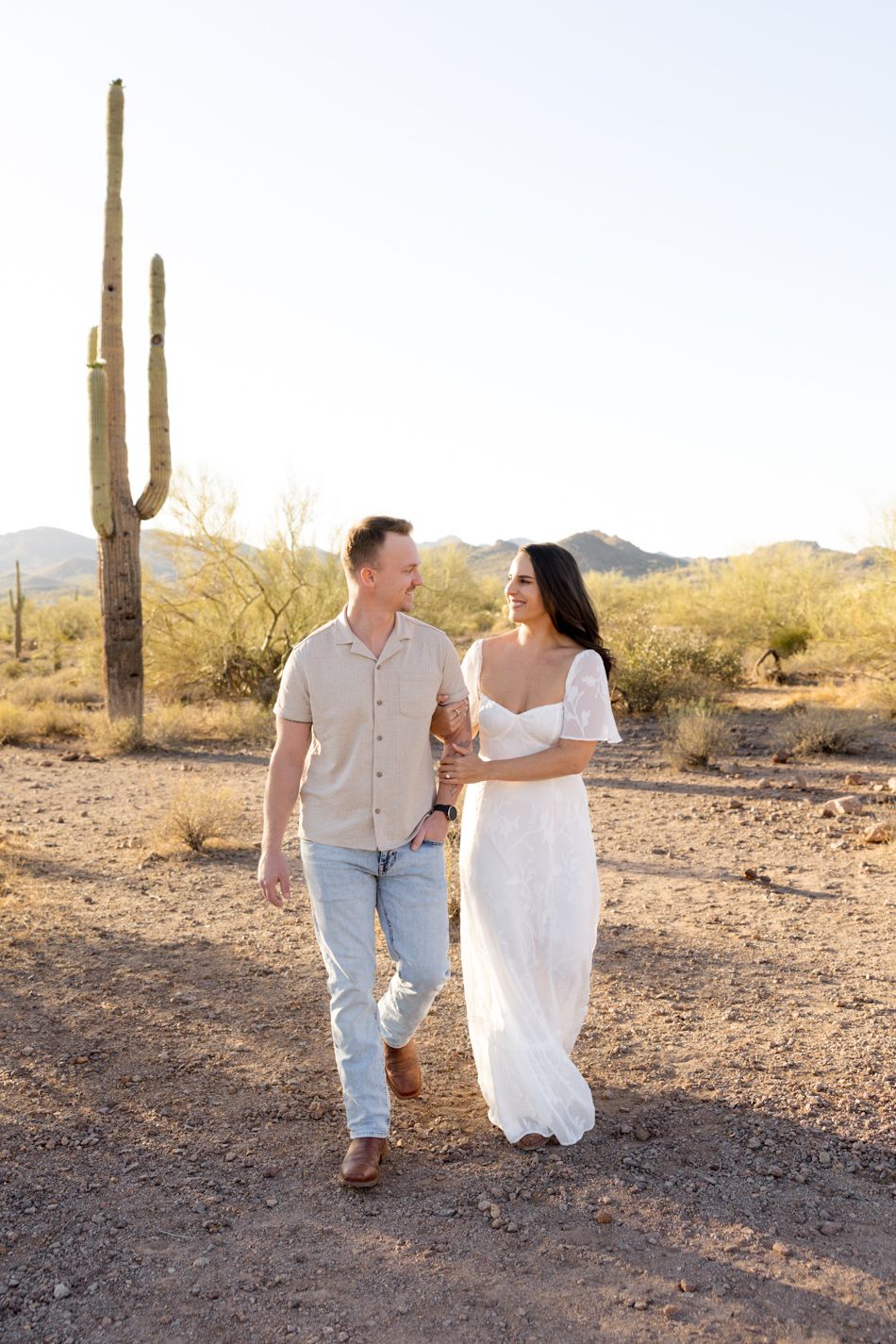 A couple walks together through a desert landscape with tall saguaro cacti and mountains in the background during sunset.