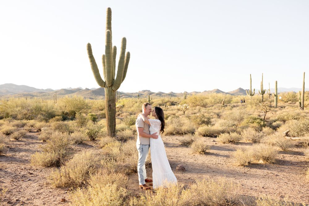 Couple embraces in a desert landscape surrounded by towering saguaro cacti and golden desert brush in Arizona.