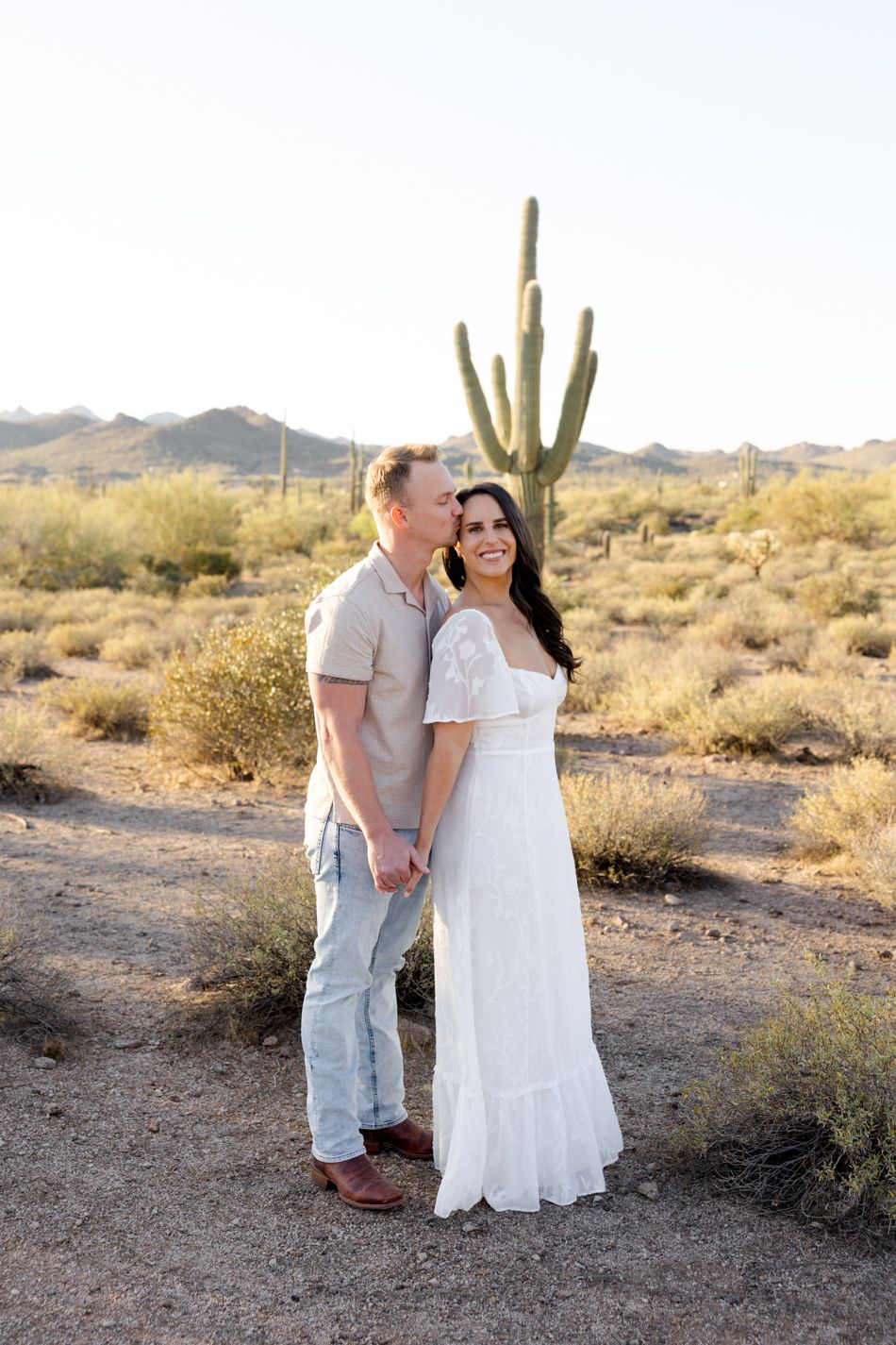 A couple poses for a romantic photo in front of a majestic saguaro cactus in the Arizona desert at sunset.