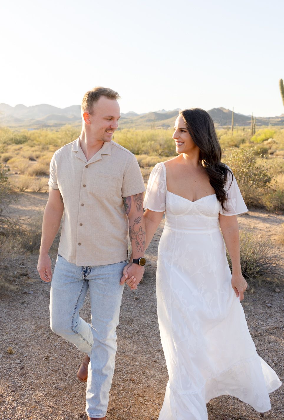 A couple holding hands while walking on a desert trail with mountains and cacti in the background at sunset.