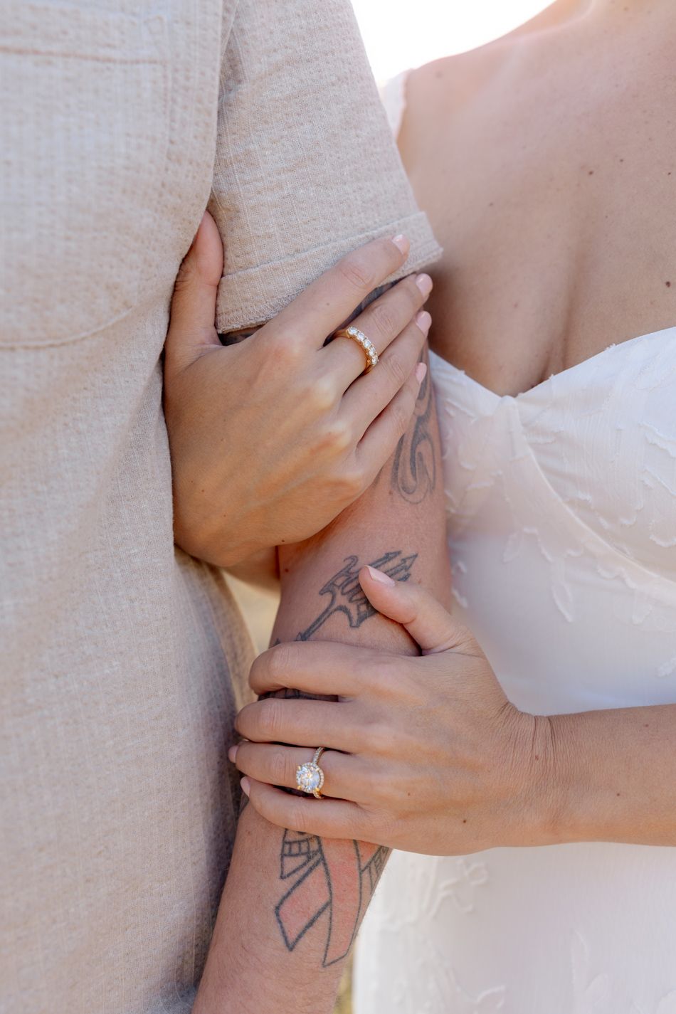 A close-up shot of hands embracing during a wedding, showing a diamond ring and arm tattoos in natural light.