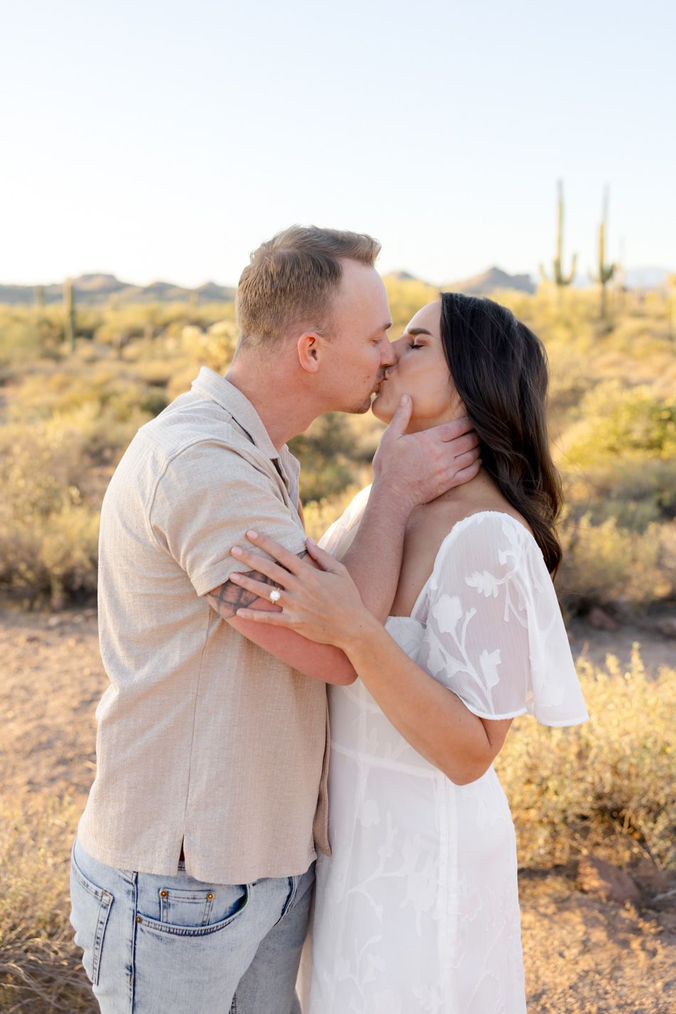 A romantic couple shares a tender kiss in the Arizona desert with saguaro cacti and golden light in the background.