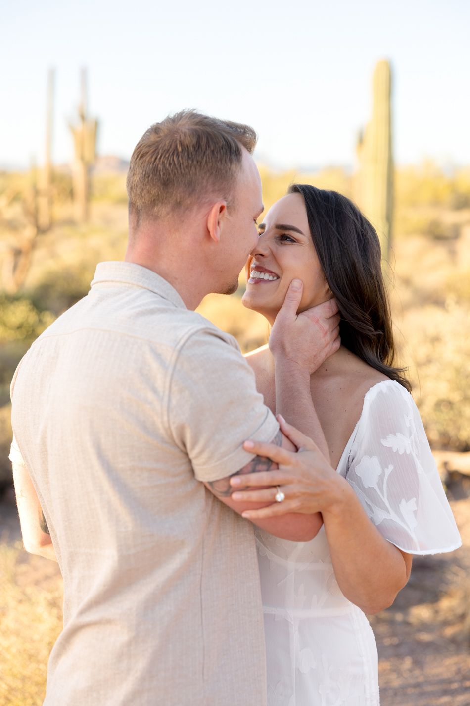 A couple shares intimate moments in a desert setting during golden hour, dressed in white against a backdrop of saguaro cacti.